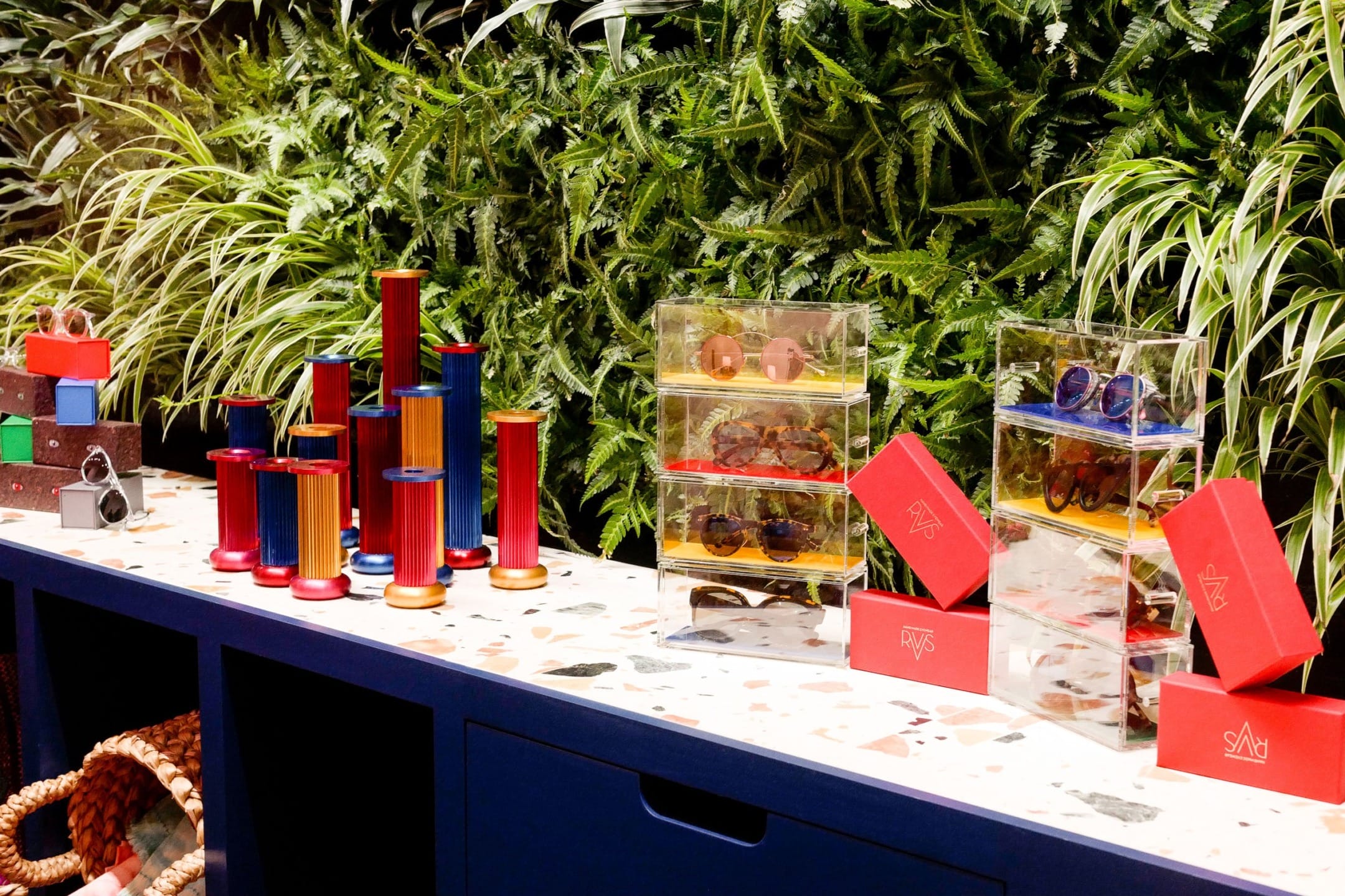 Colorful sunglasses displayed on a shelf with green plants in the background.