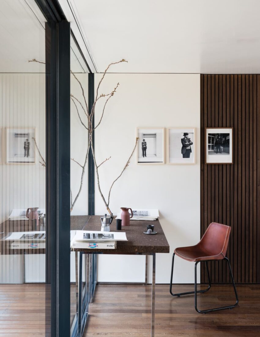 Modern home office with a sleek desk, brown leather chair, wall art, and large window looking out to a serene view.