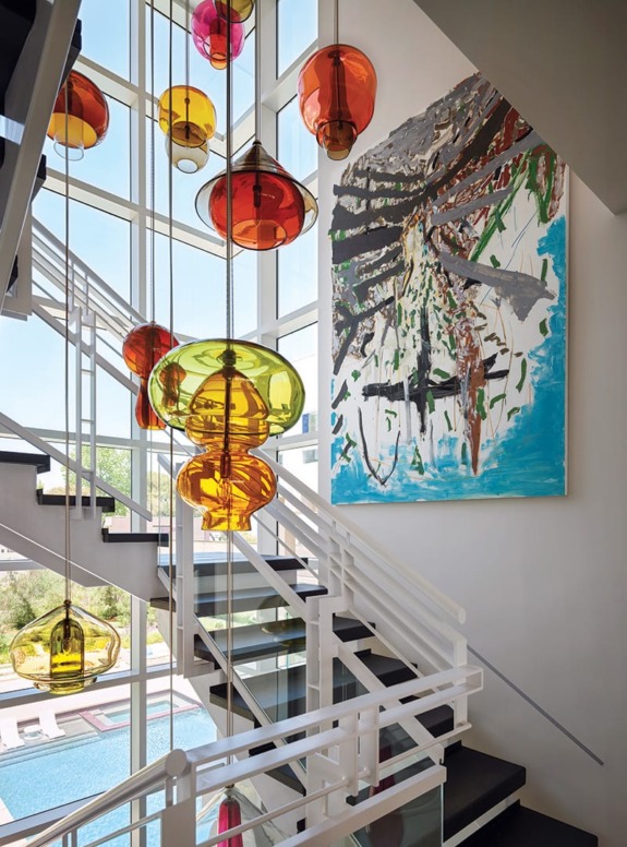 Modern staircase with colorful hanging glass lamps and abstract painting, overlooking pool and outdoor space through large windows.
