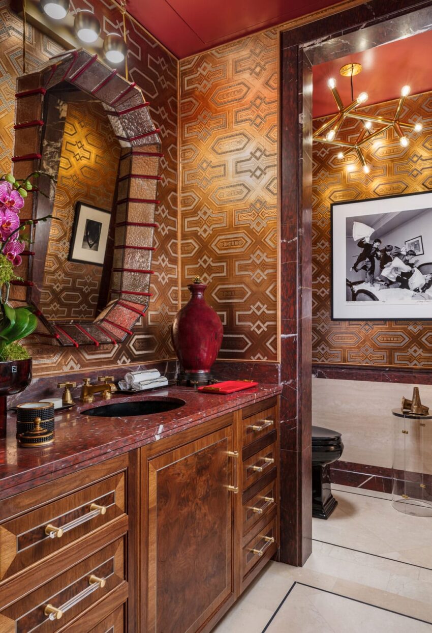 Luxurious bathroom with red and gold geometric wallpaper, wooden cabinets, ornate mirror, and decorative lighting.