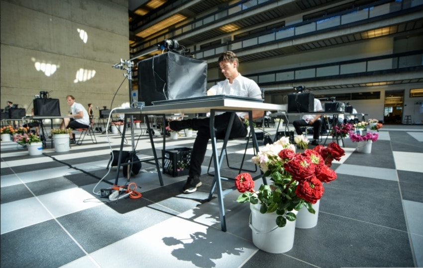Man sitting at a table with camera equipment in a spacious indoor setting, surrounded by colorful flowers in buckets.