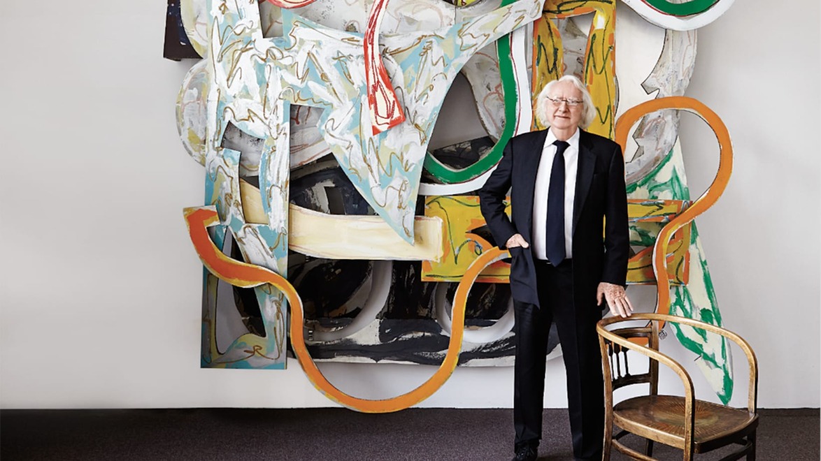 Person in a suit stands beside an abstract wall sculpture with dynamic colors and shapes, next to a wooden chair.