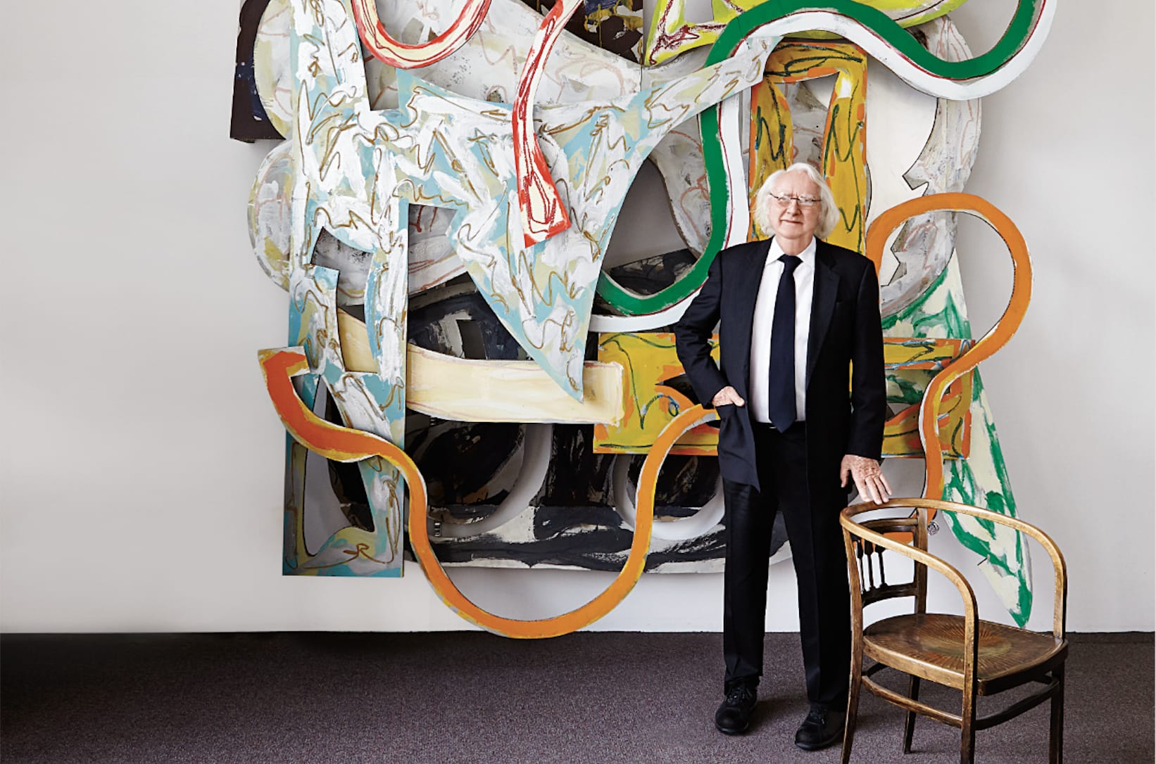 Person in a suit stands beside an abstract wall sculpture with dynamic colors and shapes, next to a wooden chair.