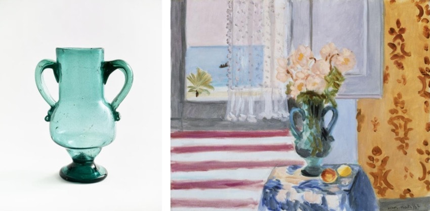 A teal glass vase with two handles next to a painting of a similar vase on a table by a window overlooking the sea.