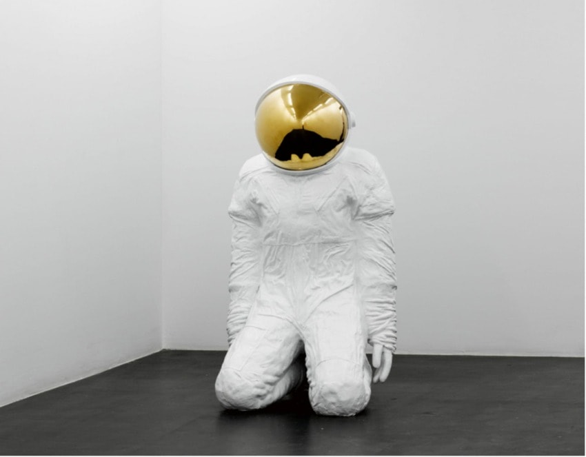 astronaut sculpture with gold helmet kneeling on black floor in a white corner room