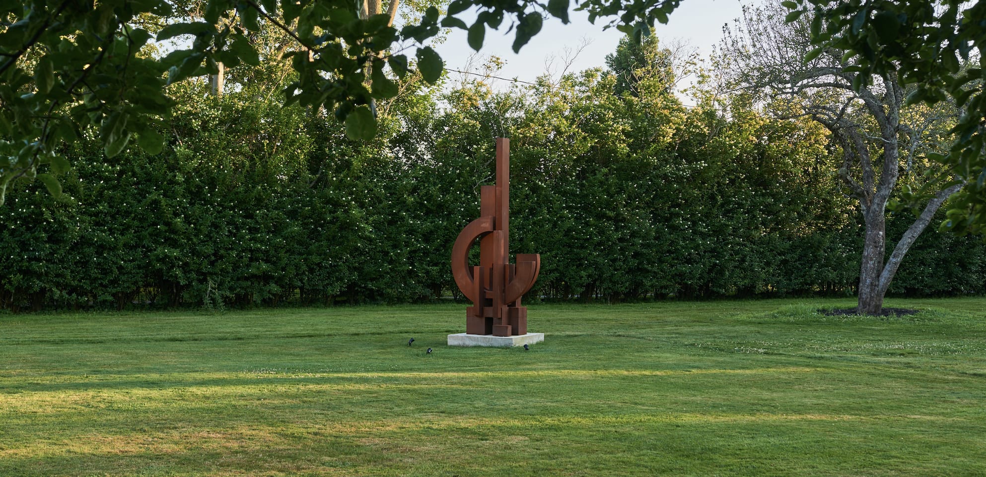 Abstract metal sculpture standing on grass with trees and bushes in the background, surrounded by a serene outdoor setting.