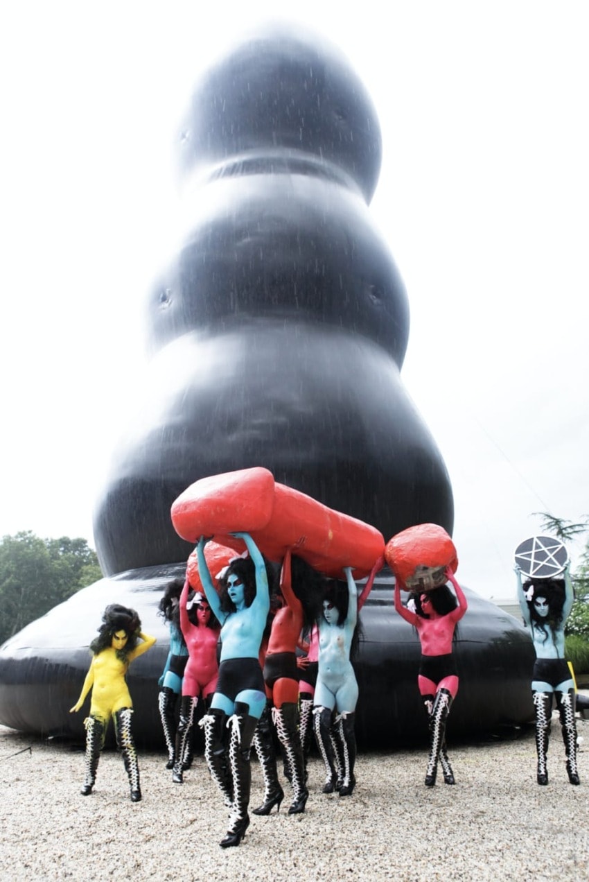 Performers in colorful body paint and black-and-white boots dancing in front of a large abstract black sculpture outdoors.