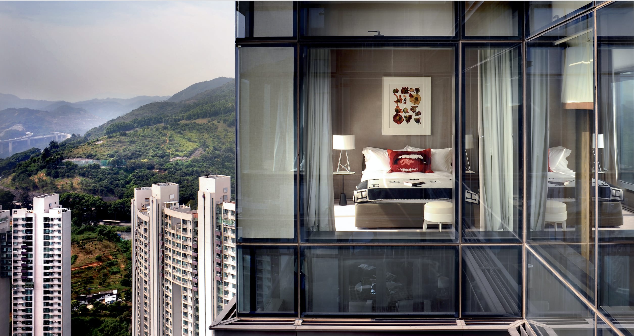 Modern hotel room with glass walls overlooking a cityscape and mountains in the background.