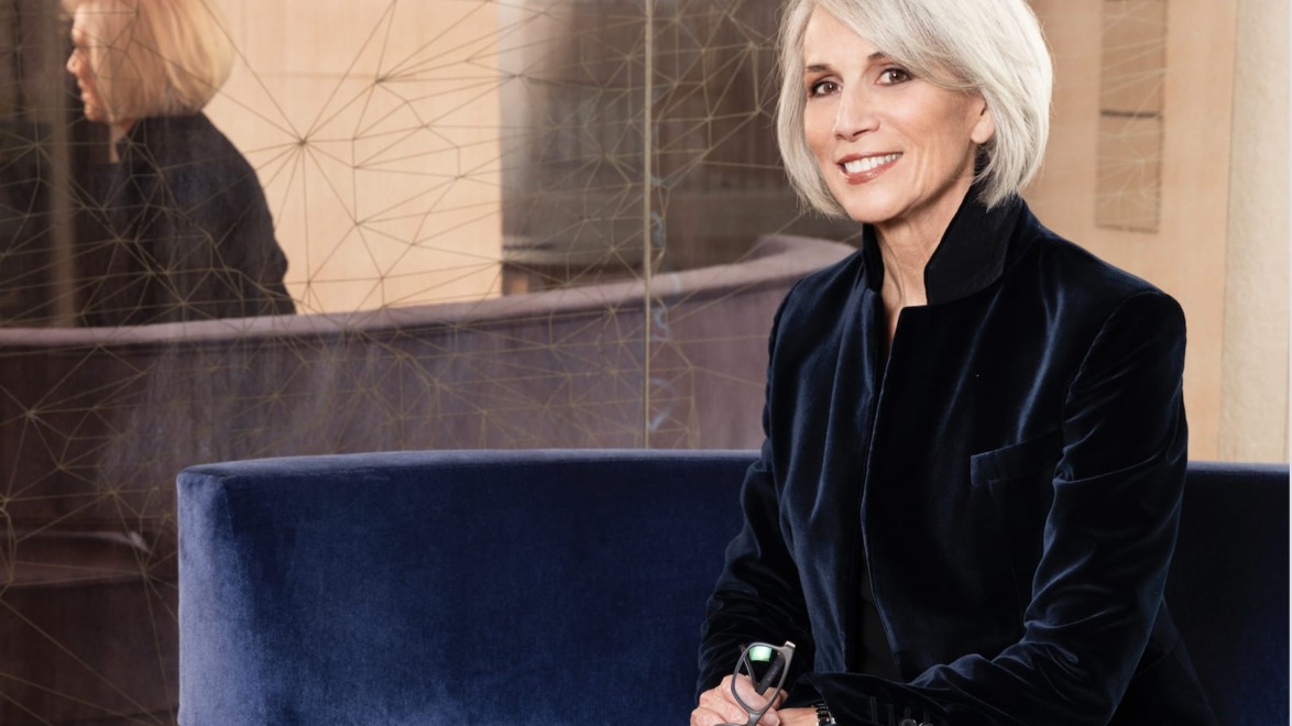 Person with short gray hair sitting on a blue couch, smiling and holding eyeglasses, reflection visible in the background.