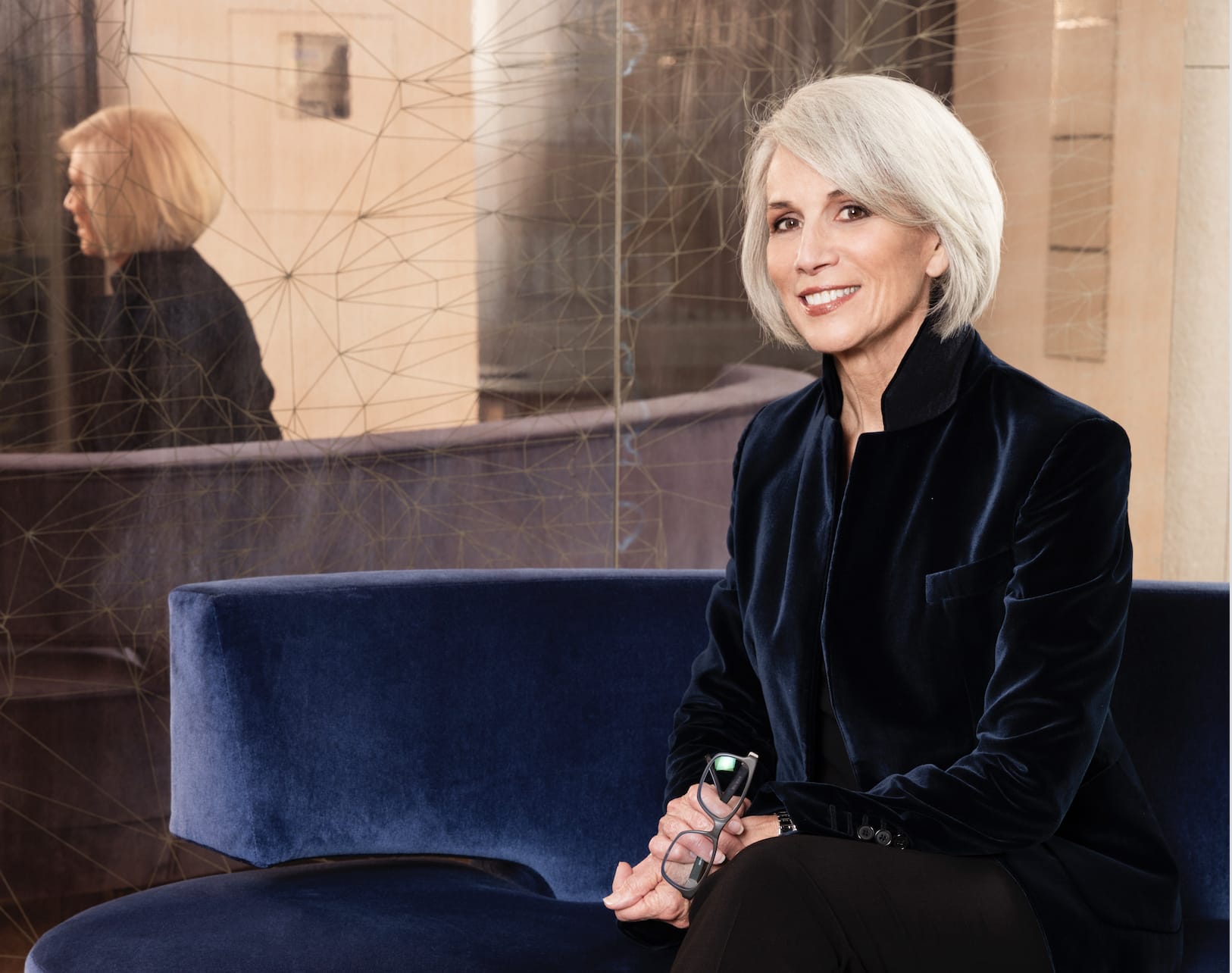 Person with short gray hair sitting on a blue couch, smiling and holding eyeglasses, reflection visible in the background.