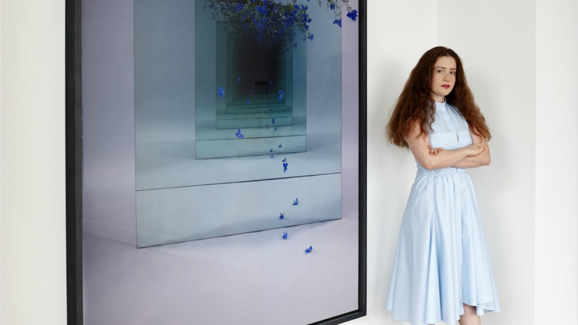Person in a light blue dress standing beside a large artwork with blue flowers on a modern, abstract background.