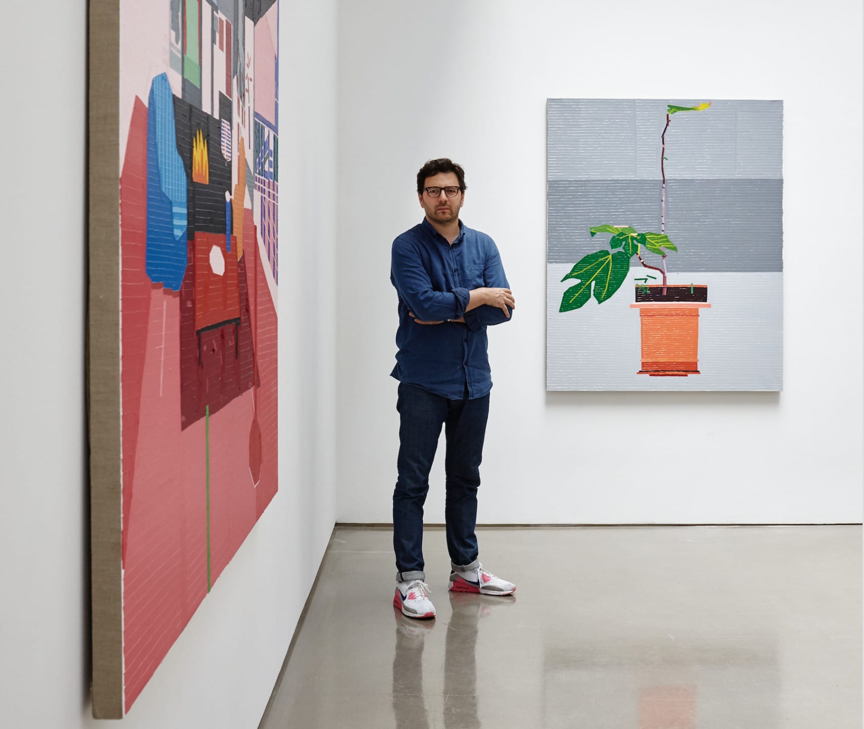 Man standing in a gallery between two colorful modern art pieces on white walls, wearing a blue shirt and jeans.