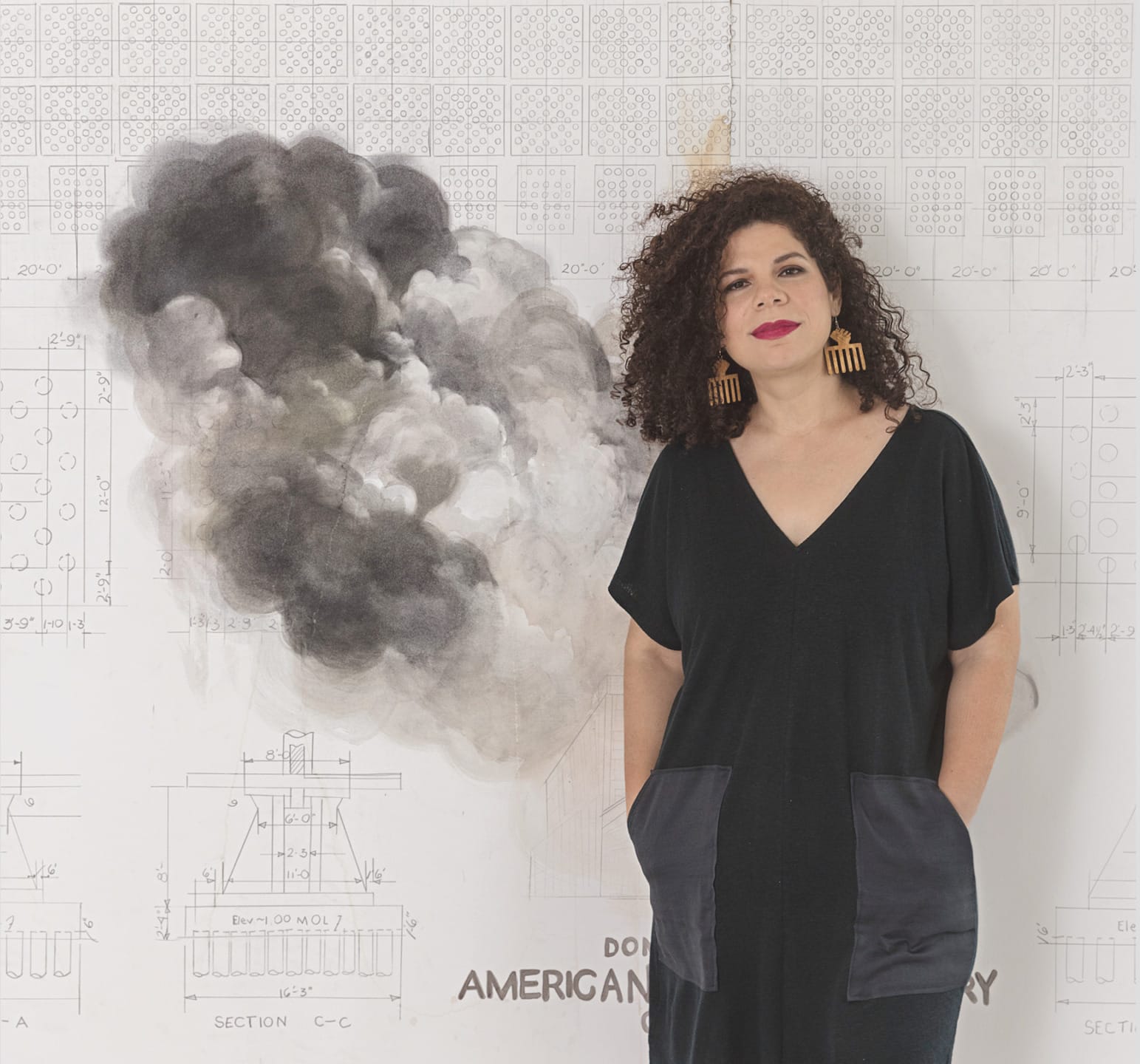 Person with curly hair standing in front of a wall with drawings and a smoke-like painting, wearing a black dress and earrings.