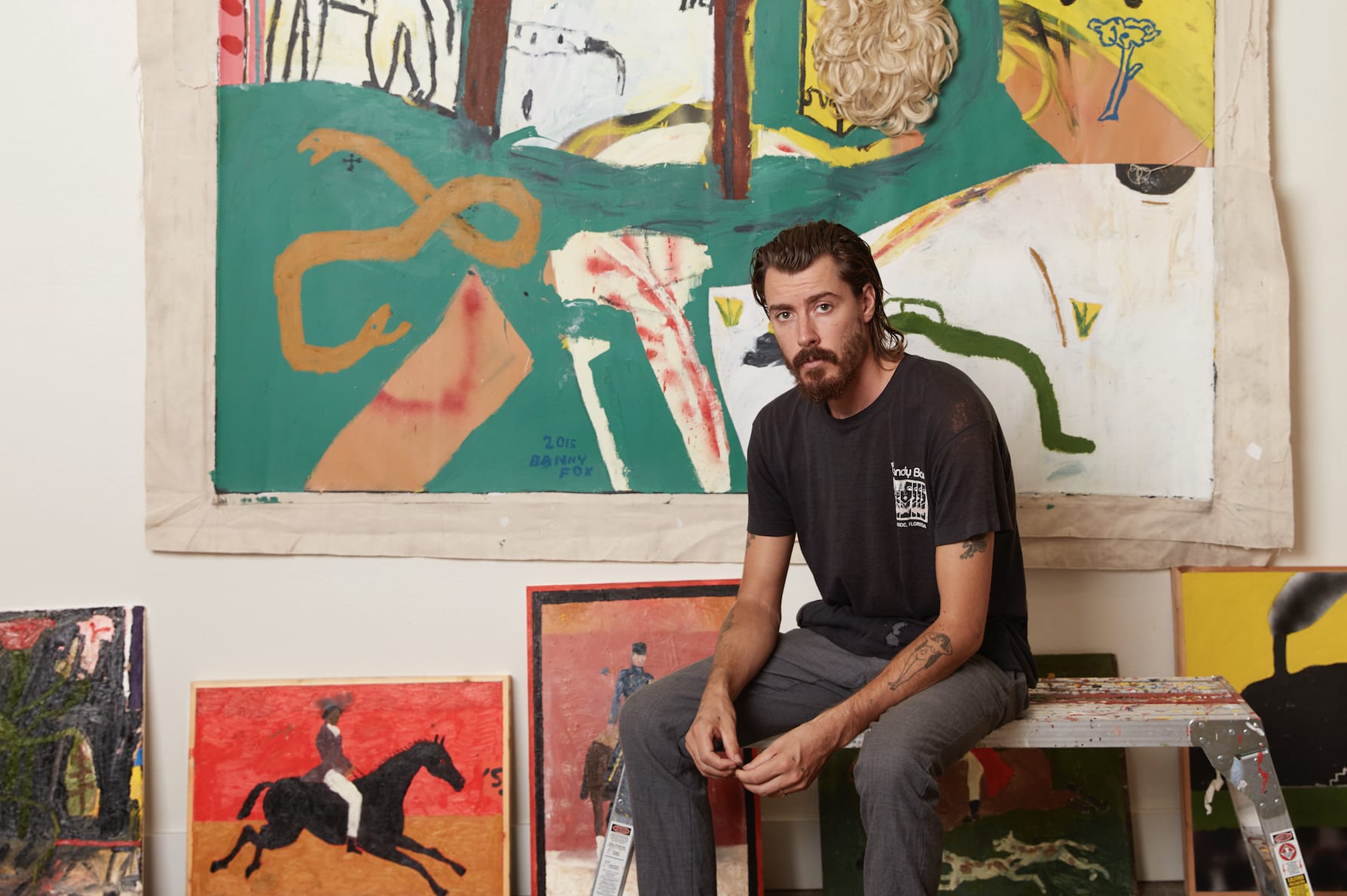 Artist sitting on a ladder in front of colorful abstract paintings in a studio setting.