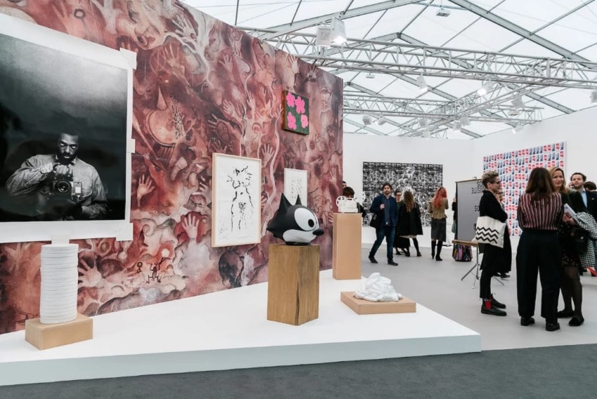 Art exhibition with various artworks displayed on walls and people viewing installations in a tented gallery space.