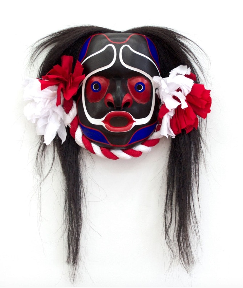Colorful ceremonial mask with red, black, and white patterns, long black hair, and red and white fabric accents.