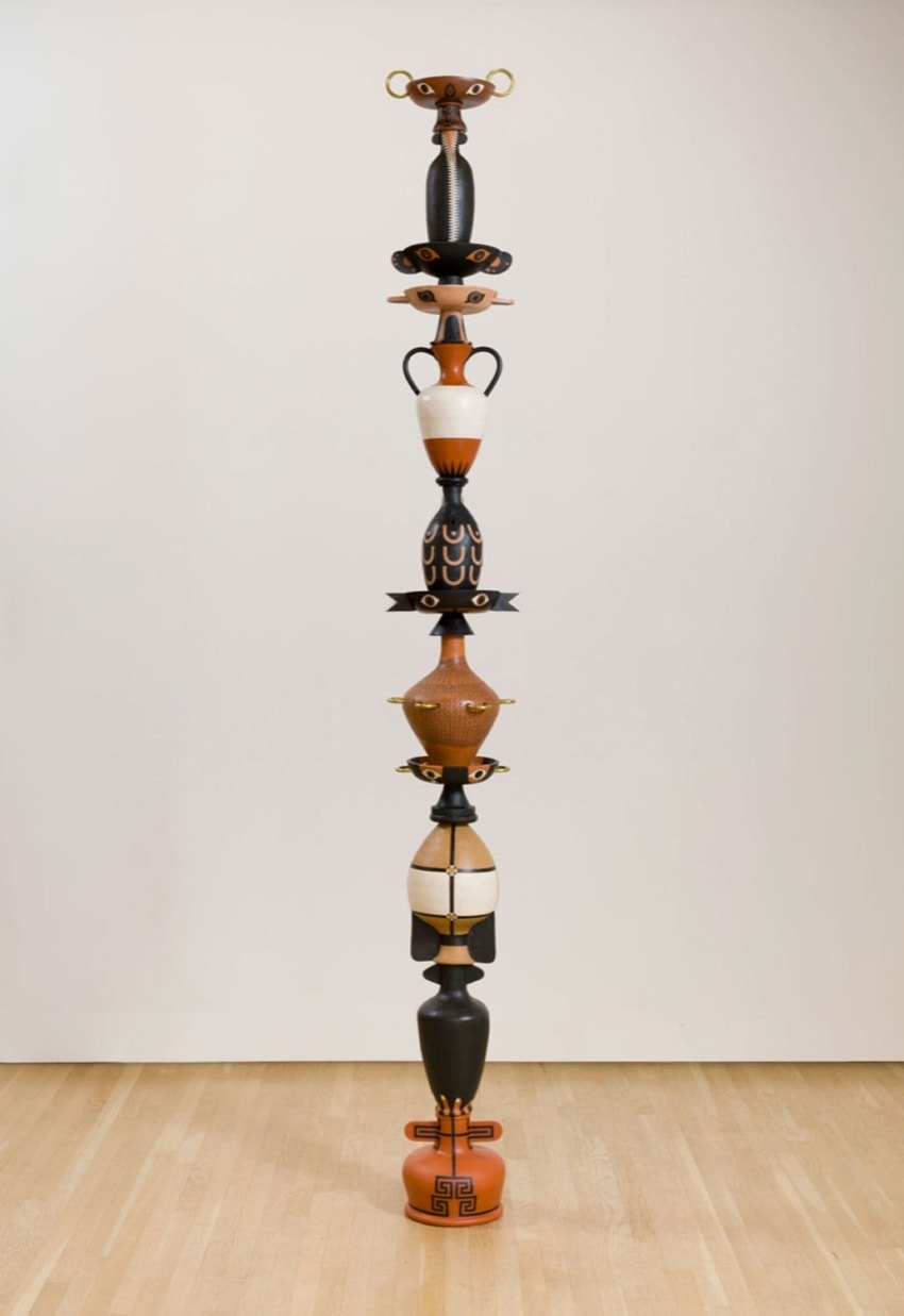 Tall totem-like sculpture made of stacked ceramic vessels in various colors and shapes on a light wood floor background.