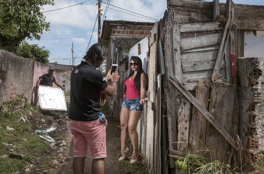 Person taking a photo of another person posing in an outdoor setting with rustic wooden fence and cloudy sky.