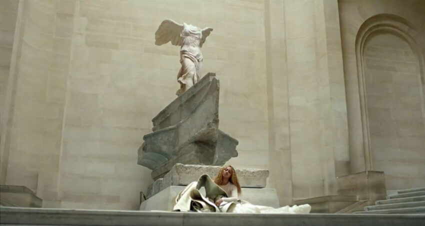 Person sitting in front of the Winged Victory of Samothrace sculpture, displayed on a large stone pedestal in a museum.