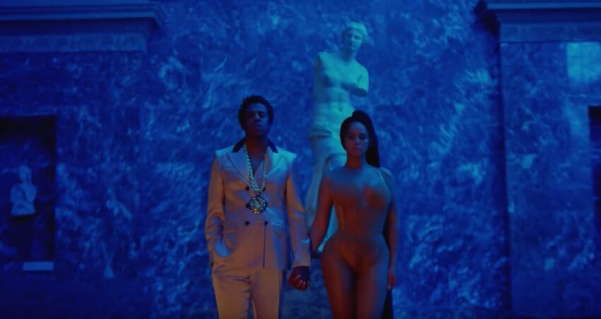 Two individuals standing in front of a blue-lit marble statue background, holding hands.