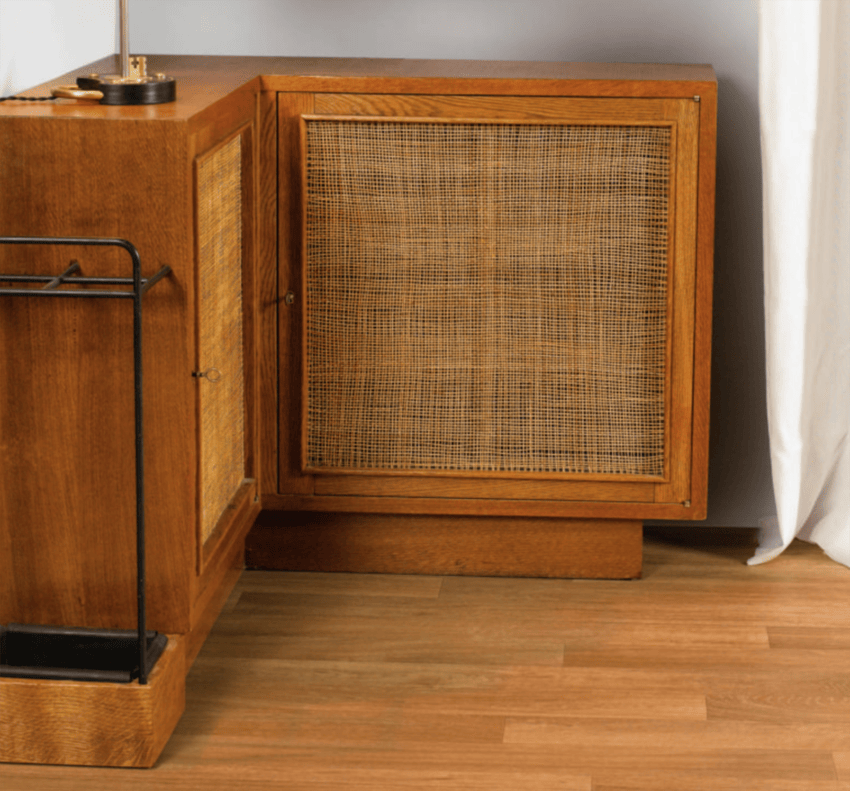 Wooden cabinet with wicker front panel and lamp on top, next to a white curtain on a wooden floor.