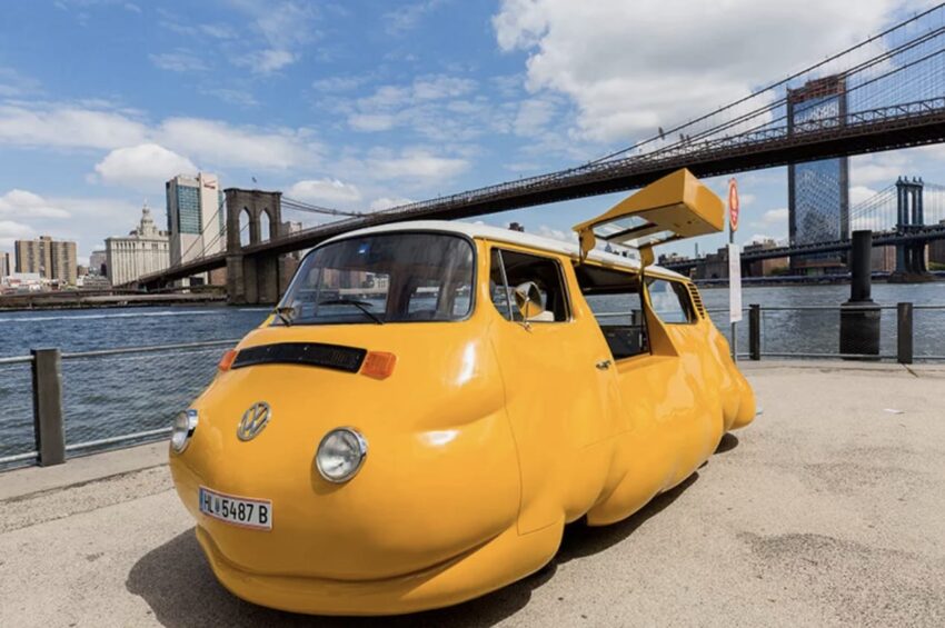 Yellow stretched Volkswagen Beetle limo parked near water with city skyline and bridge in the background.