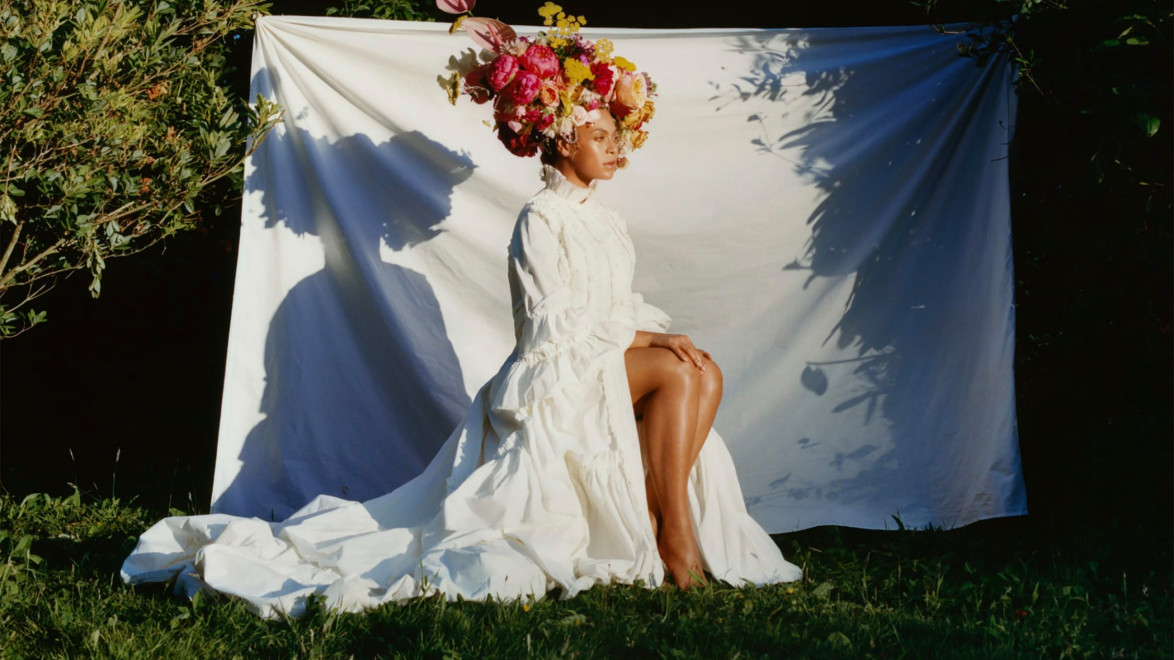 Person in a white dress with a colorful flower headdress sitting outdoors against a white backdrop.