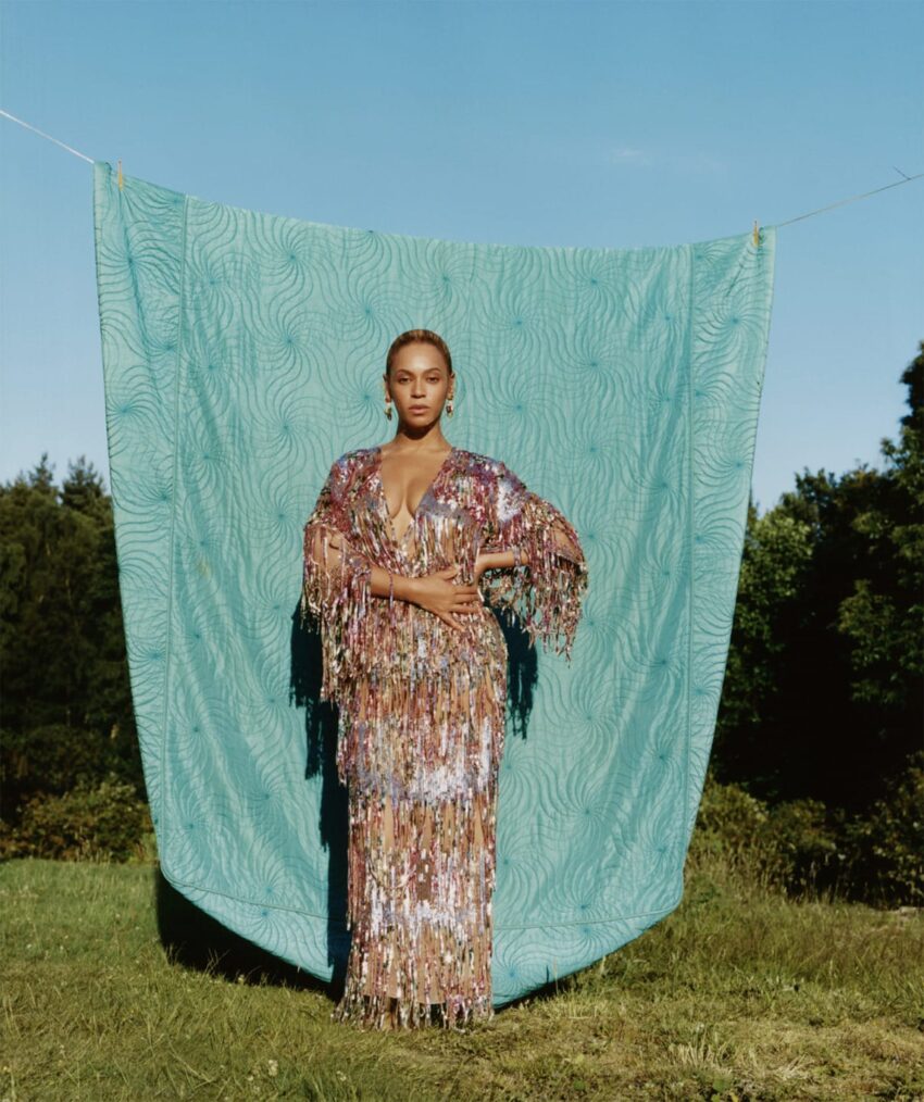Person wearing a colorful sequined dress stands in front of a turquoise patterned fabric outdoors.