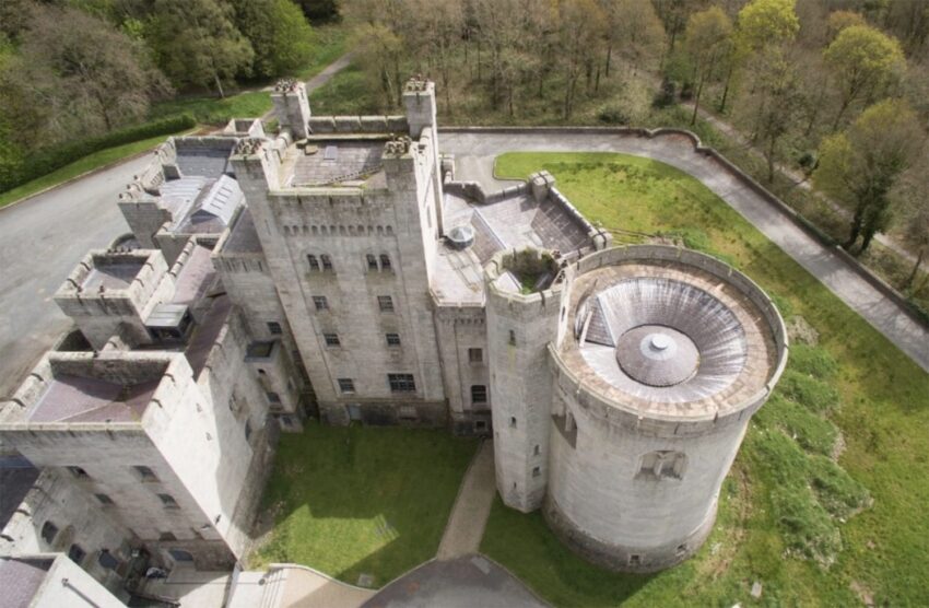 Aerial view of an old stone castle surrounded by trees and a circular tower in the foreground.