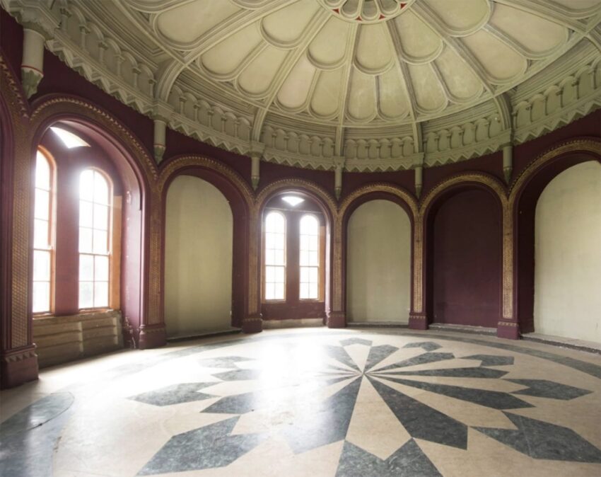 Interior of an empty circular room with a patterned floor, arched windows, and a decorative domed ceiling.