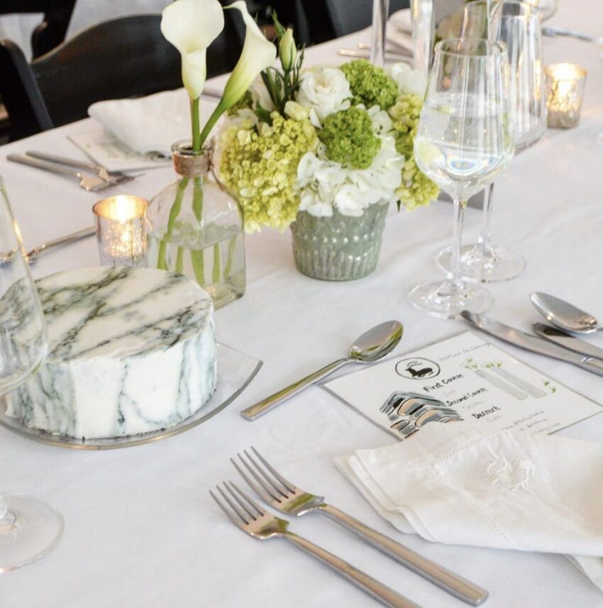 Elegant table setting with a floral centerpiece, marble cheese wheel, cutlery, wine glasses, and a menu on a white tablecloth.