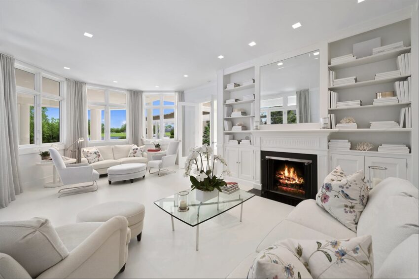 Modern living room with white decor, a glass coffee table, and a fireplace, surrounded by large windows and built-in shelves.