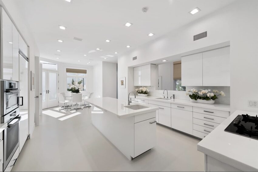 Modern white kitchen with sleek cabinets, island with sink, and recessed lighting in a bright open space.