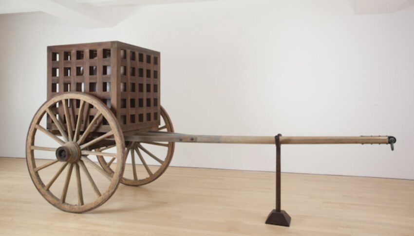 Wooden wheeled cart with a lattice box structure displayed in a minimalist gallery setting.