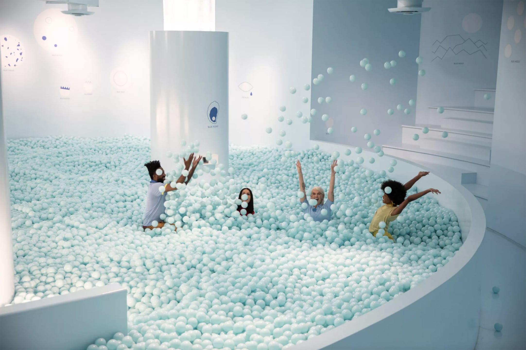 People playing in a large indoor ball pit filled with light blue balls, with white walls and stairs in the background.