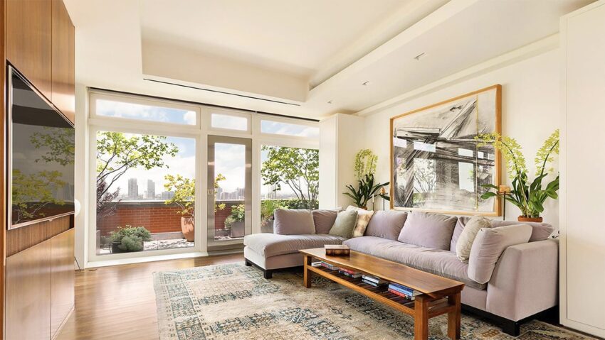 Modern living room with large windows, cozy grey sectional sofa, wooden coffee table, and greenery outside.