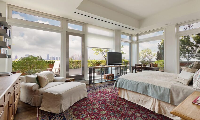 Spacious bedroom with floor-to-ceiling windows, city skyline view, cozy seating, and elegant decor.