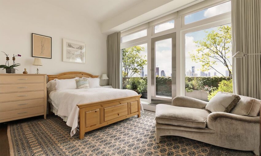 Bright bedroom with large windows, city view, comfy chair, wooden bed, dresser, and patterned rug.