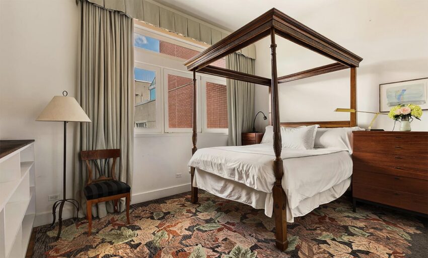 Spacious bedroom with a four-poster wooden bed, floral carpet, window with curtains, dresser, chair, and lamp.