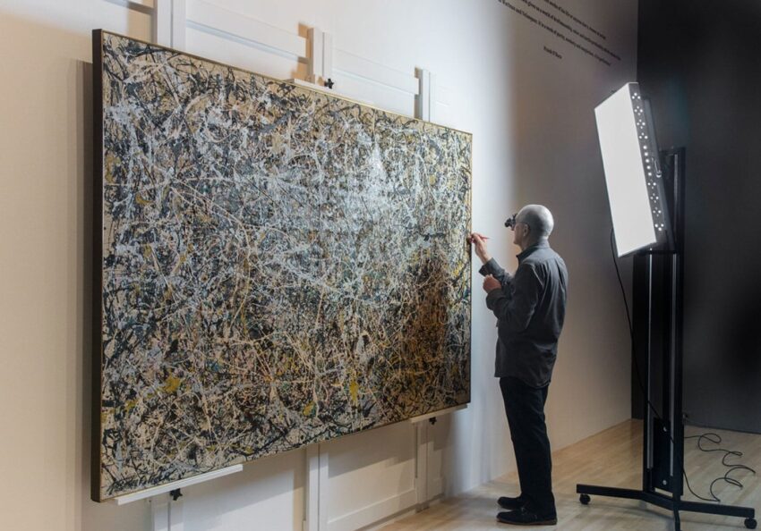 Person closely examining a large abstract painting in an art gallery setting with professional lighting equipment nearby.