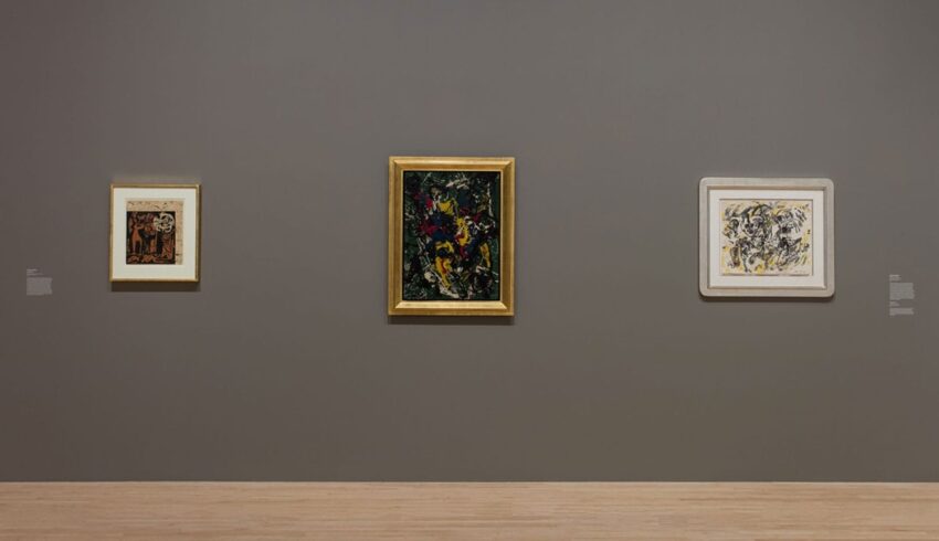 Three framed abstract paintings displayed on a museum wall with wooden flooring.