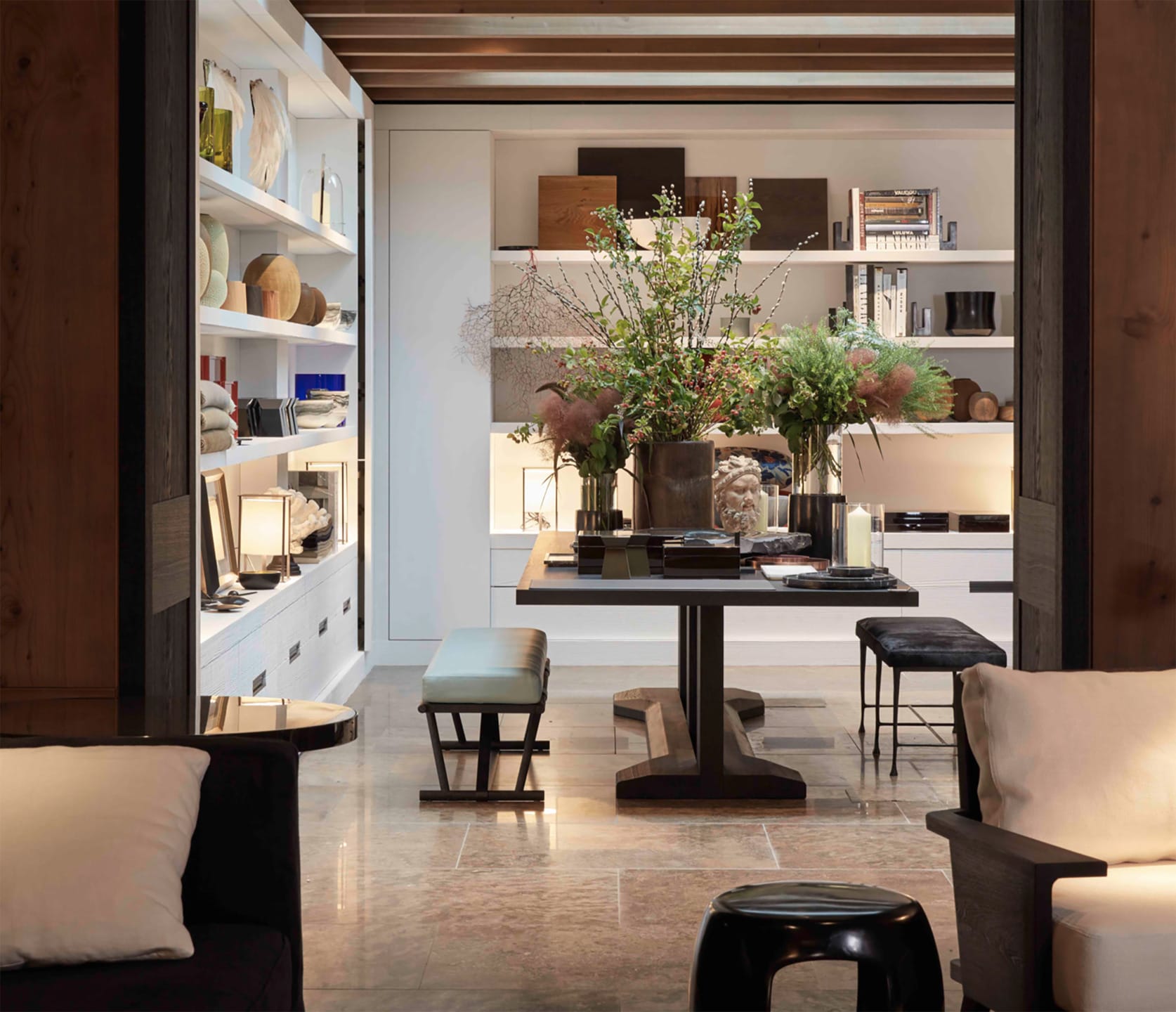 Modern living room interior with natural decor, wood ceiling, and shelves displaying books and vases, centered table with plants.