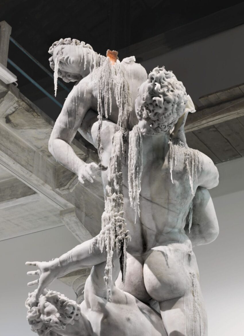 Marble sculpture of two intertwined figures, partially covered in dripped candle wax, displayed in a modern gallery setting.
