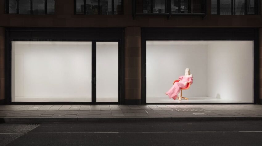 Person seated in a bright pink outfit inside a well-lit, minimalistic display window next to a darkened empty window.
