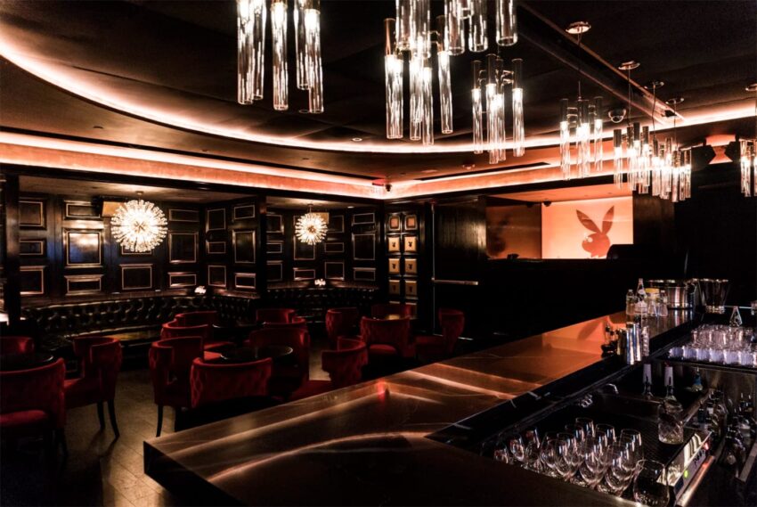 Dimly lit lounge with chandelier lights, plush seating, and a bar area featuring a bunny logo on the back wall.