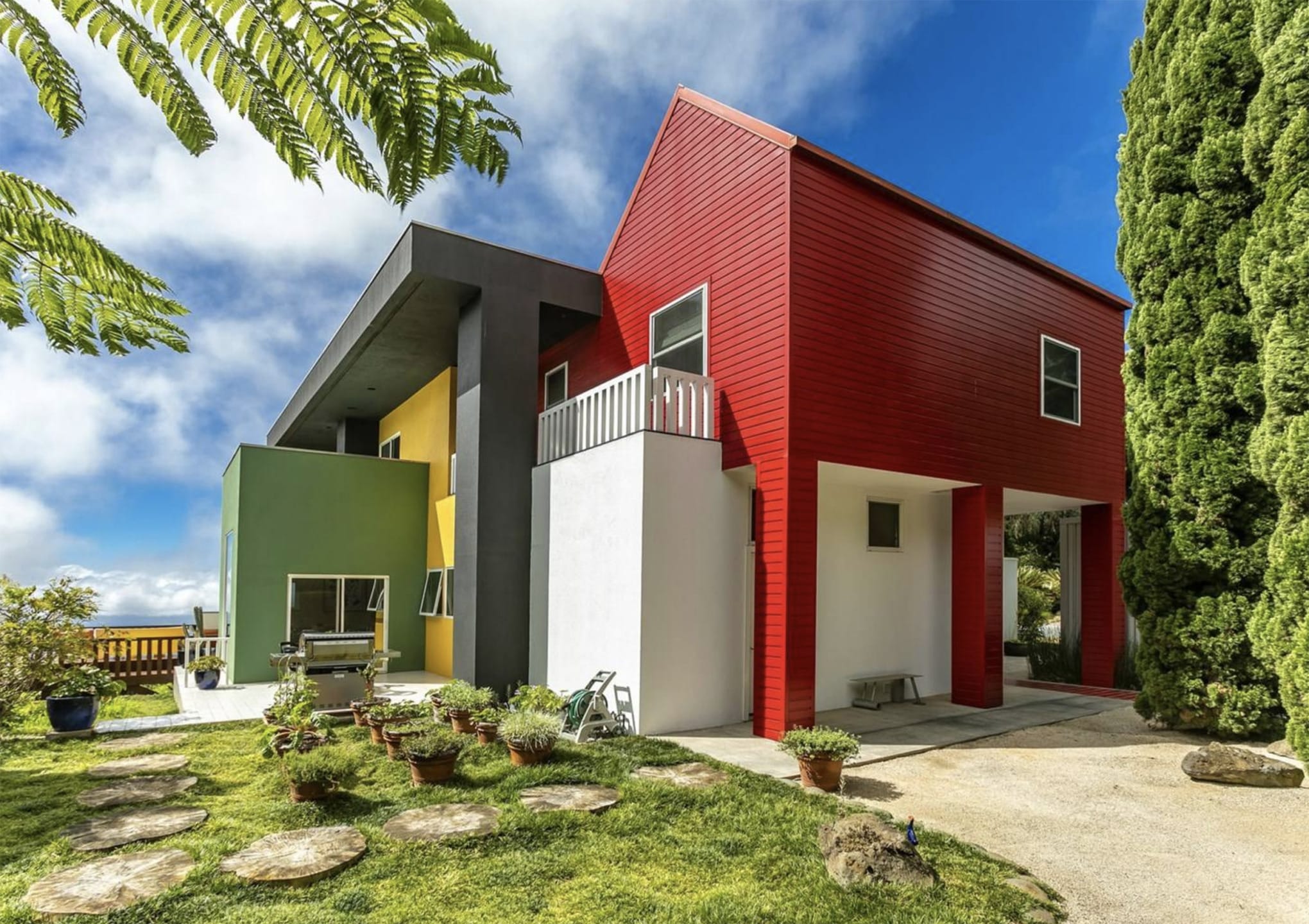 Modern colorful house with red, green, and yellow sections, surrounded by a garden and trees under a blue sky.