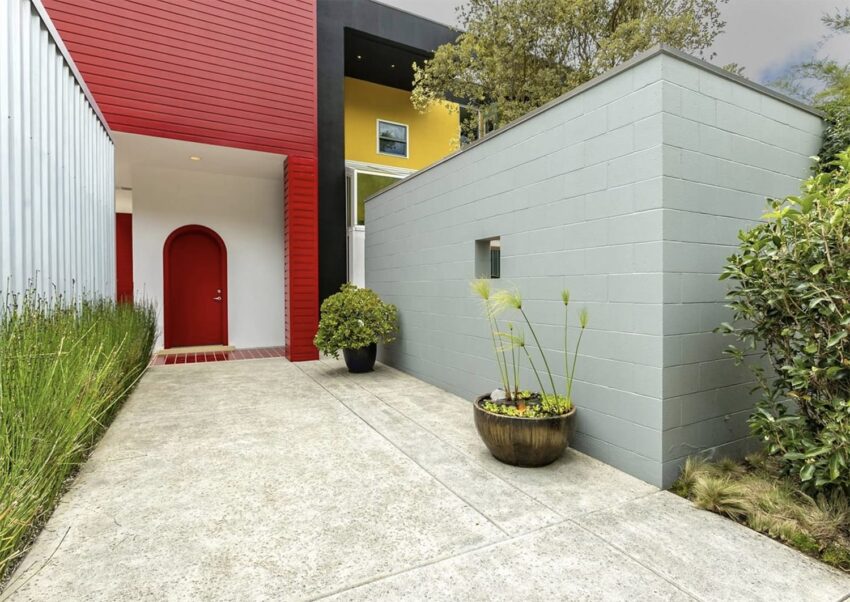 Modern house entrance with bright red door, gray wall, and potted plants surrounded by greenery.