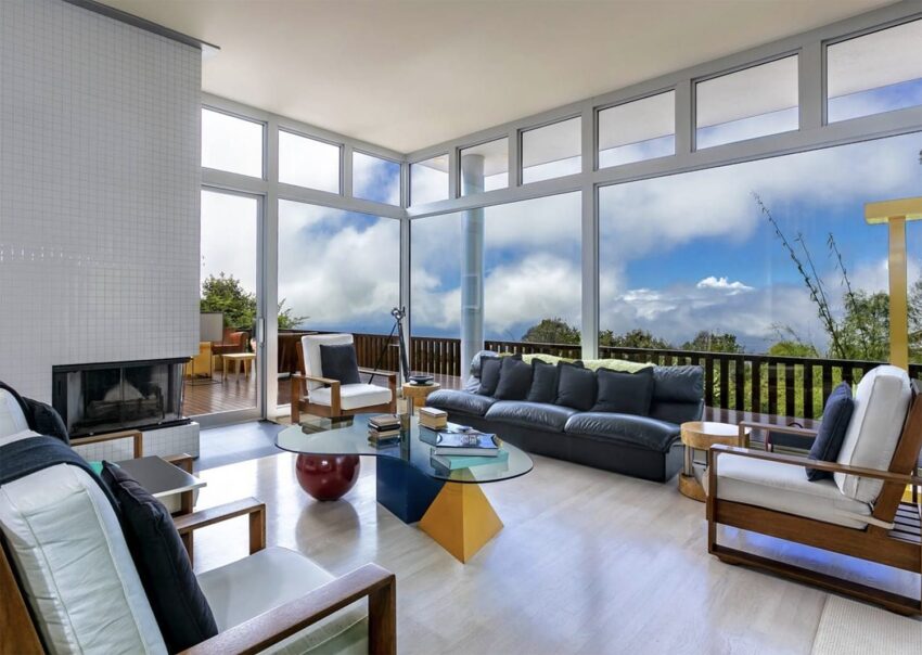 Modern living room with large windows, black sofa, glass coffee table, and view of clouds and greenery outside.