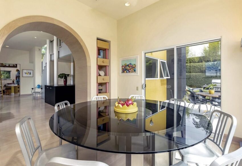 Modern dining room with a round glass table, metal chairs, and an open archway leading to a bright living area.