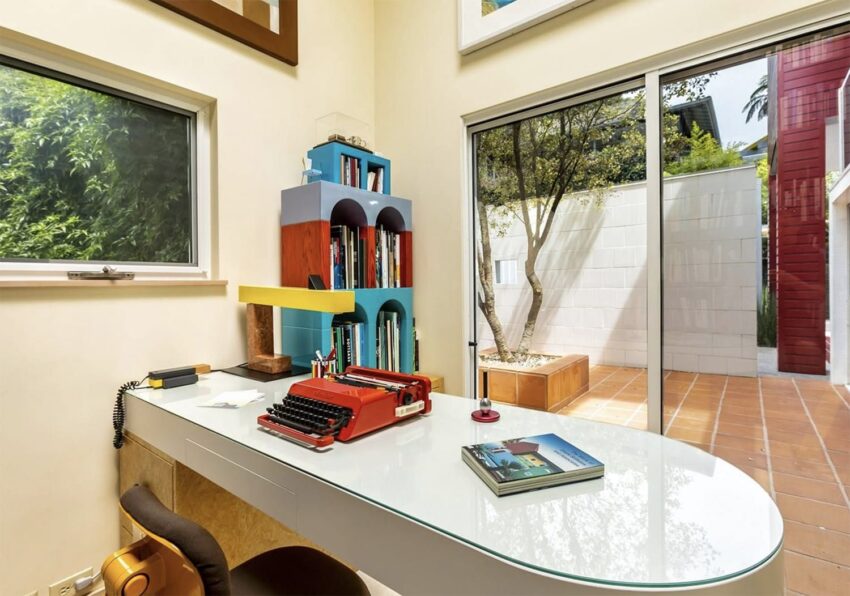 Modern home office with white desk, vintage red typewriter, and large windows overlooking a patio and garden.